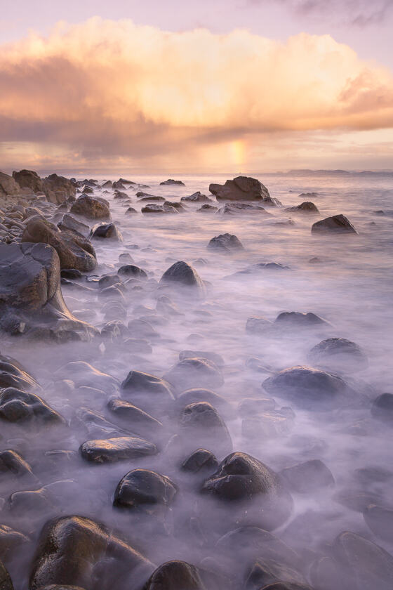 A long exposure of the sea lapping at a boulder-strewn shore on the Ross of Mull, with a rainbow visible in the direction of mainland Scotland.
